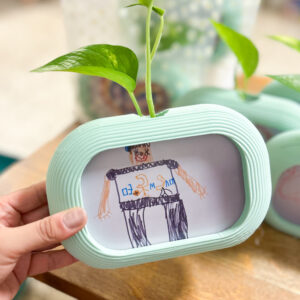 ייחור ירוק של פוטוס שצומח מתוך מסגרת תלת־ממדית בצבע תכלת עם ציור של ילד. מסגרת דו צדדית לילדים עם ציור צבעוני של ילד וייחור ירוק של פוטוס